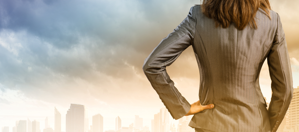 waist up view of a woman wearing a business suit facing away from the camera and staring out toward a foggy city. 