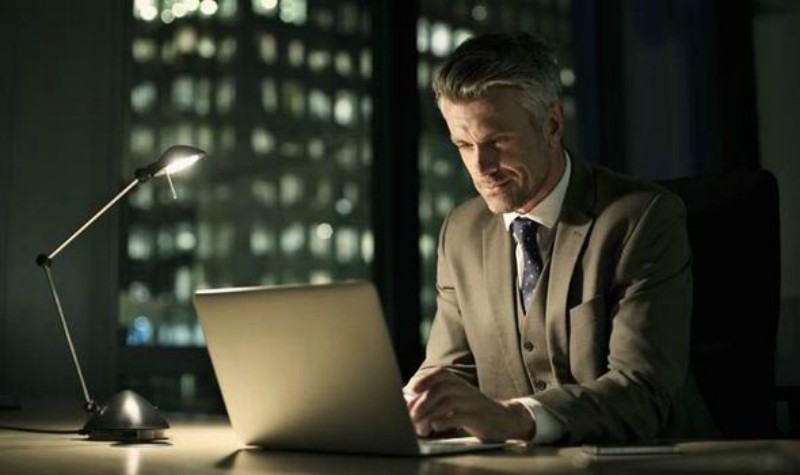 Man sitting at a dimly lit desk looking at a laptop.