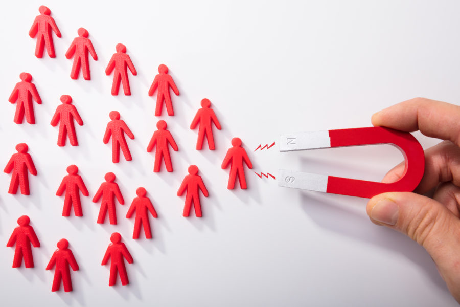 Tiny red magnets shaped like people grouped in a triangle form with a hand holding a horseshoe shaped magnet trying to attract the people.